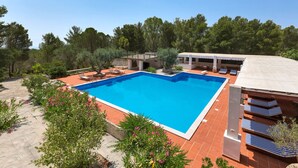 Una piscina rectangular con solárium azulejado, rodeada de árboles y una zona de estar exterior cubierta con sillones de descanso.