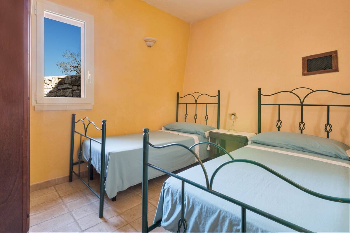 Una habitación con dos camas, un ventanal que da a un árbol y un muro de piedra, y una lámpara fijada a la pared.