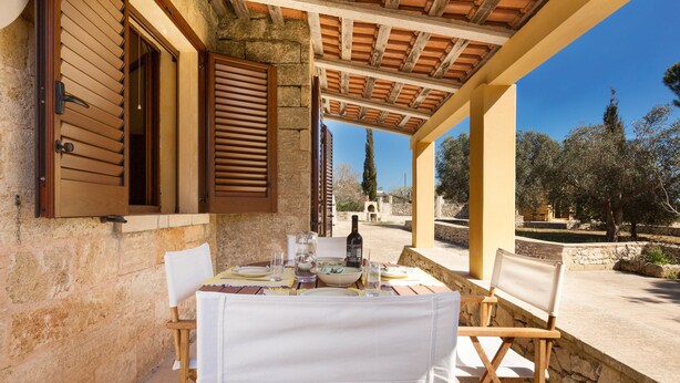 Un espacio de comedor al aire libre cubierto, con pared de piedra, contraventanas de madera y una mesa preparada para dos.