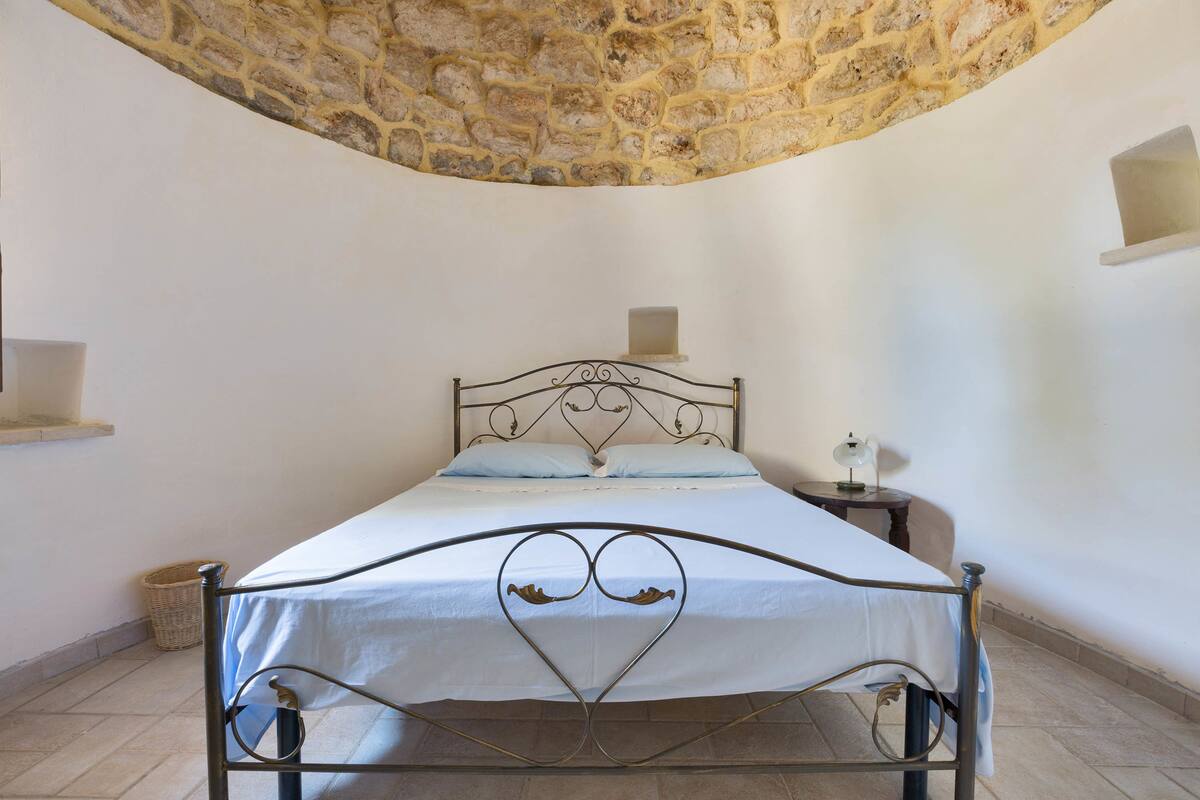 Una cama con estructura metálica y diseño en forma de corazón, una pared de piedra y una mesita de noche con lámpara.