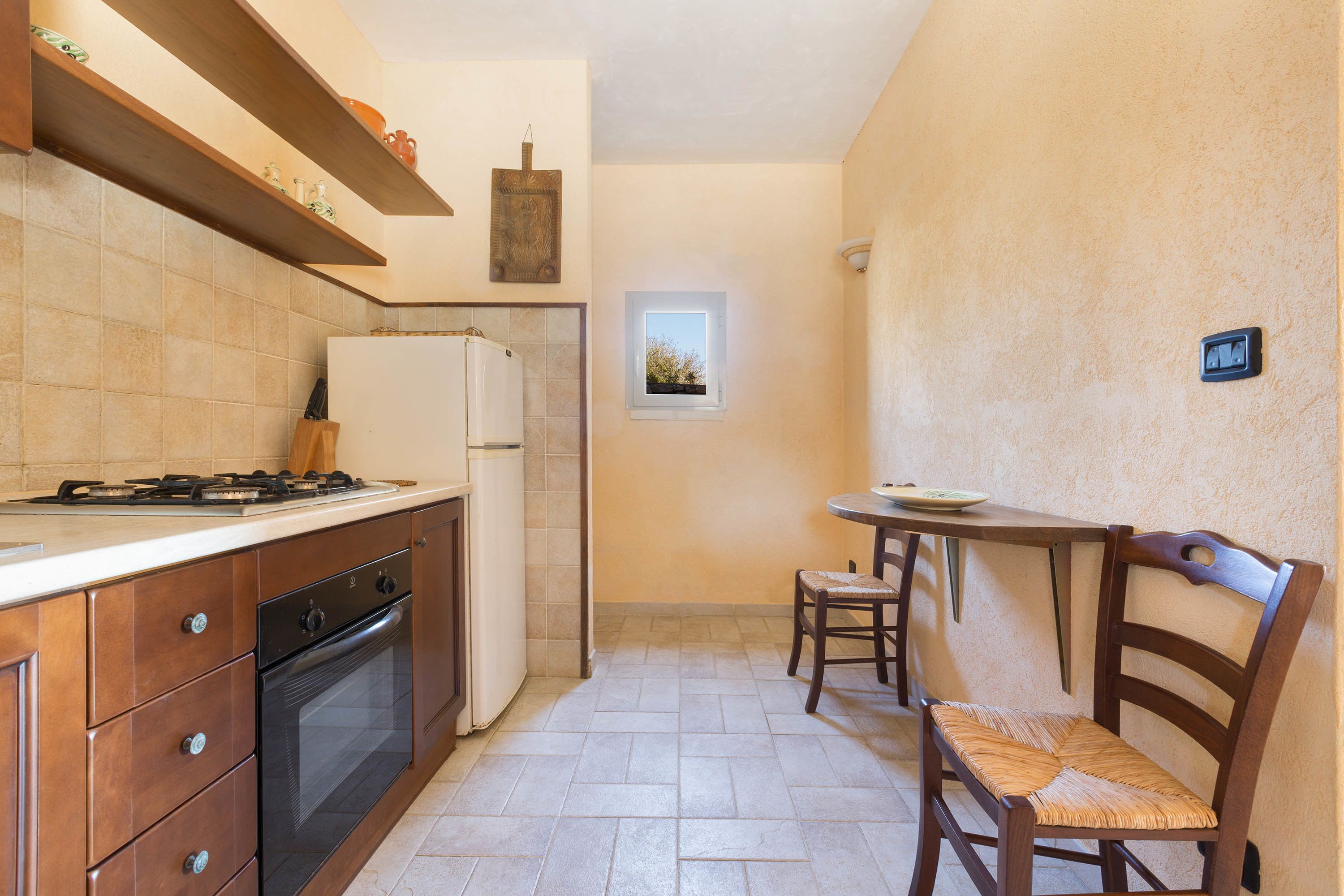 Eine kompakte Küche mit Gasherd, Backofen und Kühlschrank. Ein kleiner Esstisch mit zwei Stühlen steht darunter, über ihm befindet sich ein Fenster.