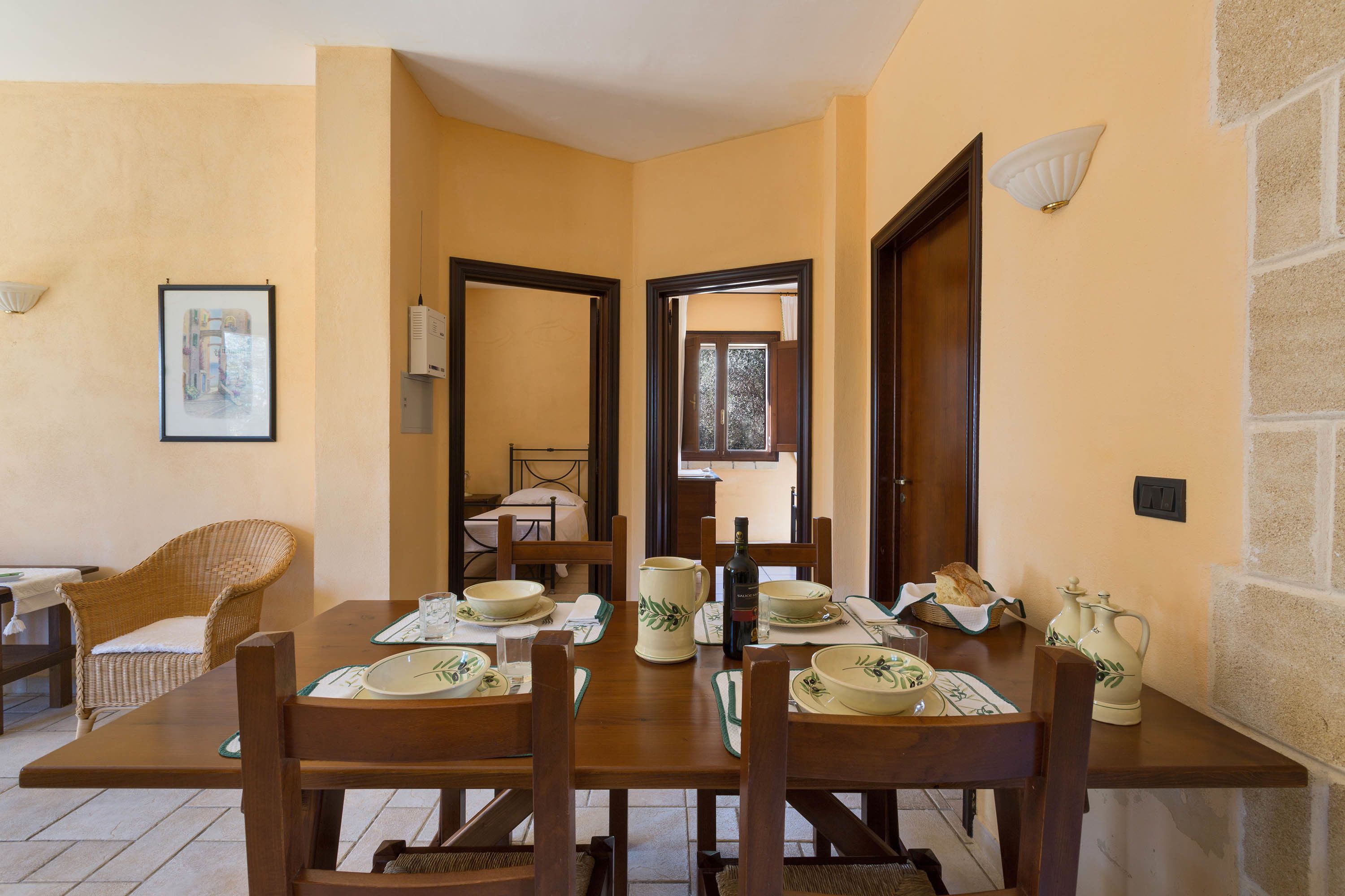 A dining area with a wooden table set for four, surrounded by chairs. There are two doors leading to other rooms, a framed picture on the wall, and a light fixture above the table.