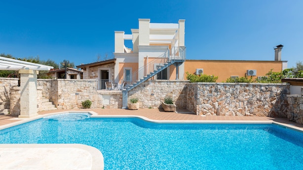 Une piscine bordée d’un mur en pierre et un escalier menant à un bâtiment blanc.