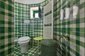 A bathroom with green and white checkered tiles, a white toilet, a small green stool, and a wicker basket. - Trulli Il Cappero (Ostuni)