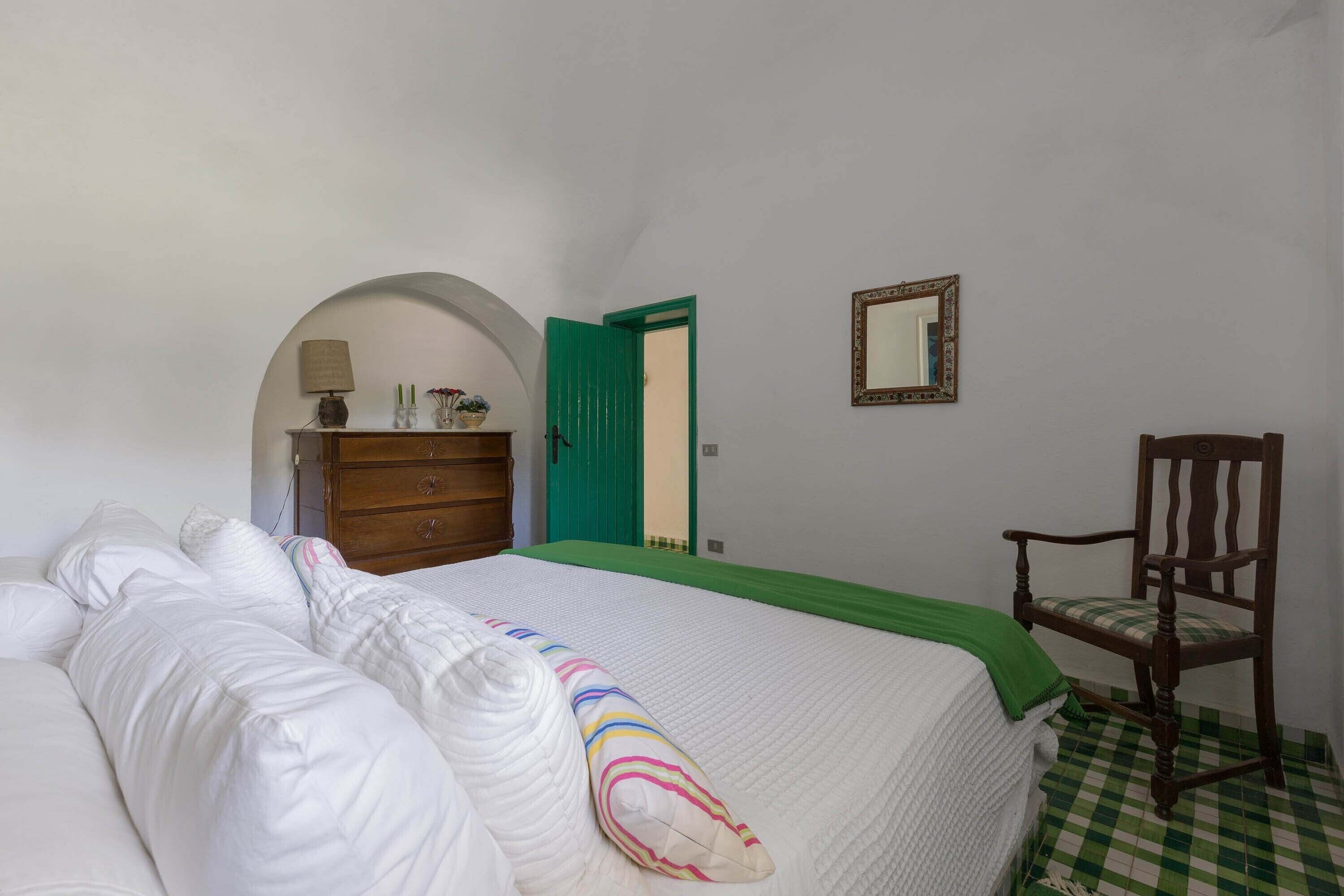 A bedroom with a bed, a wooden dresser, a chair, a mirror, and a green door.