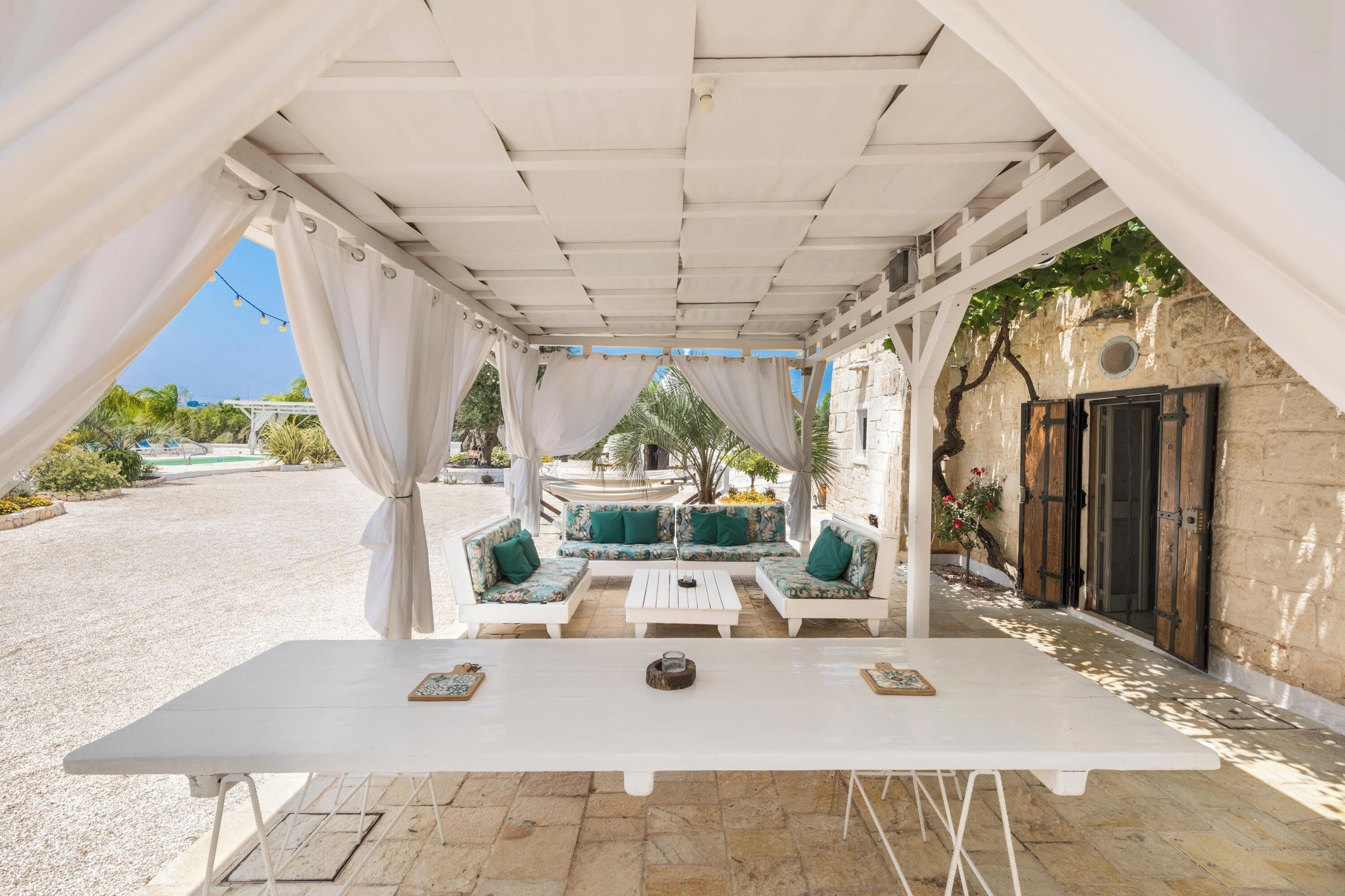 An outdoor seating area with white tables and green cushioned sofas, a pergola overhead, and a view of a garden and building in the background.