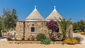 A stone building with two conical roofs, surrounded by various plants and trees. - Trulli Ion (Ostuni)