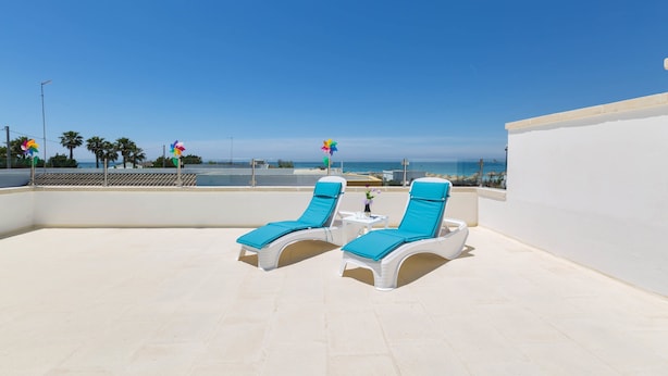 Eine Dachterrasse mit zwei weißen Liegestühlen mit blauen Kissen, einem kleinen Tisch mit Blumenvase und freiem Blick auf das Meer und Palmen im Hintergrund.
