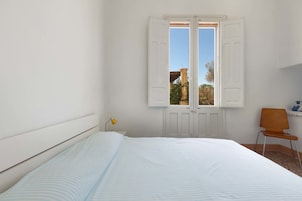 Quarto com cama, janela com persianas, uma cadeira e uma mesinha.
