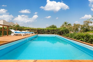 Une piscine rectangulaire au fond carrelé, entourée d’une terrasse en bois. On y trouve des chaises longues avec des coussins bleus et une structure en forme de pergola.
