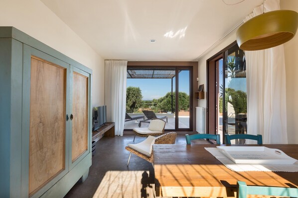 Una habitación moderna con una mesa de madera, sillas de mimbre, un televisor y vistas al exterior.