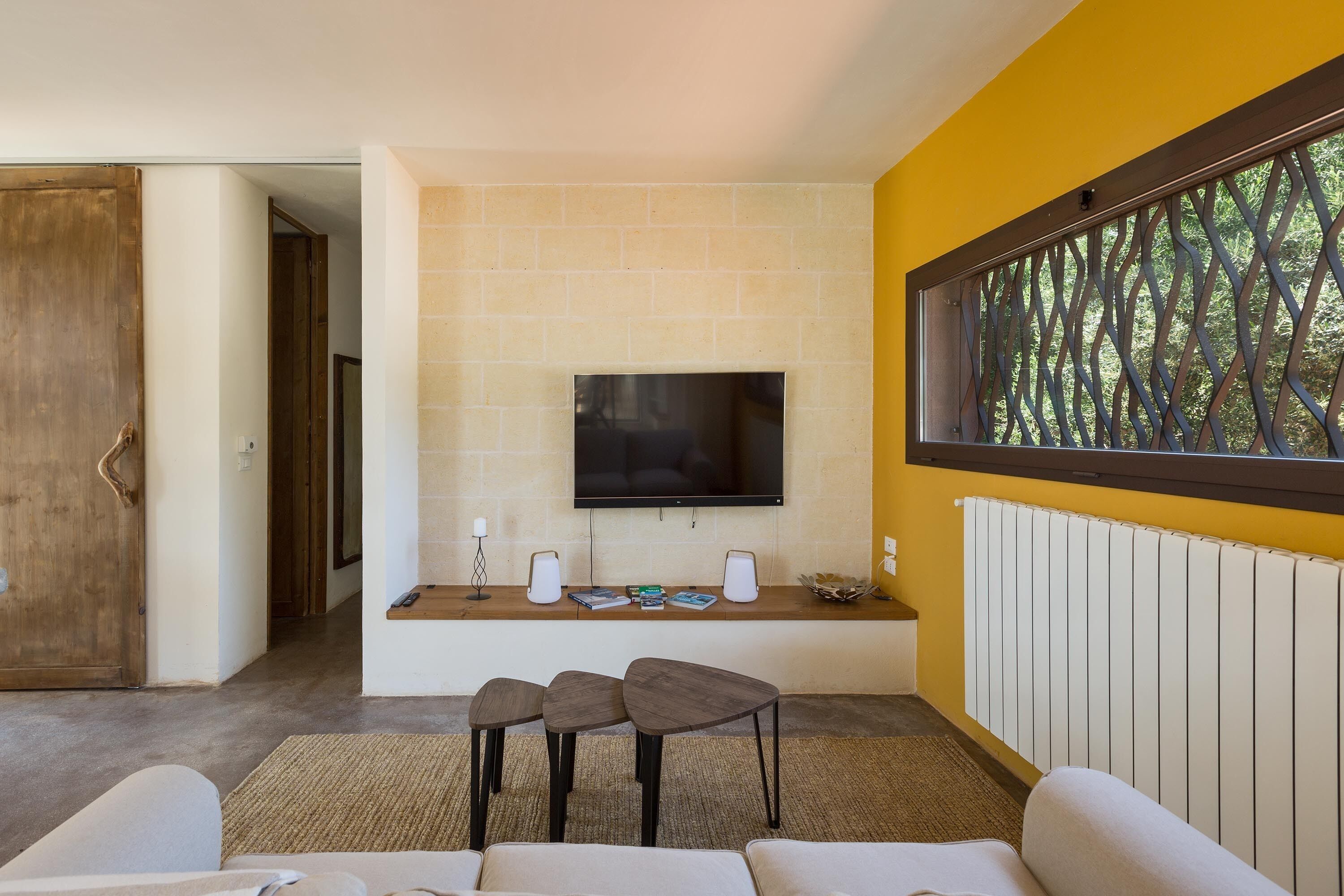 A modern living room with a TV mounted on a brick wall, a wooden door, and a large window with a view of trees.
