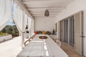 A long dining table with chairs, white curtains, a hanging lantern, and a view of the outdoors.