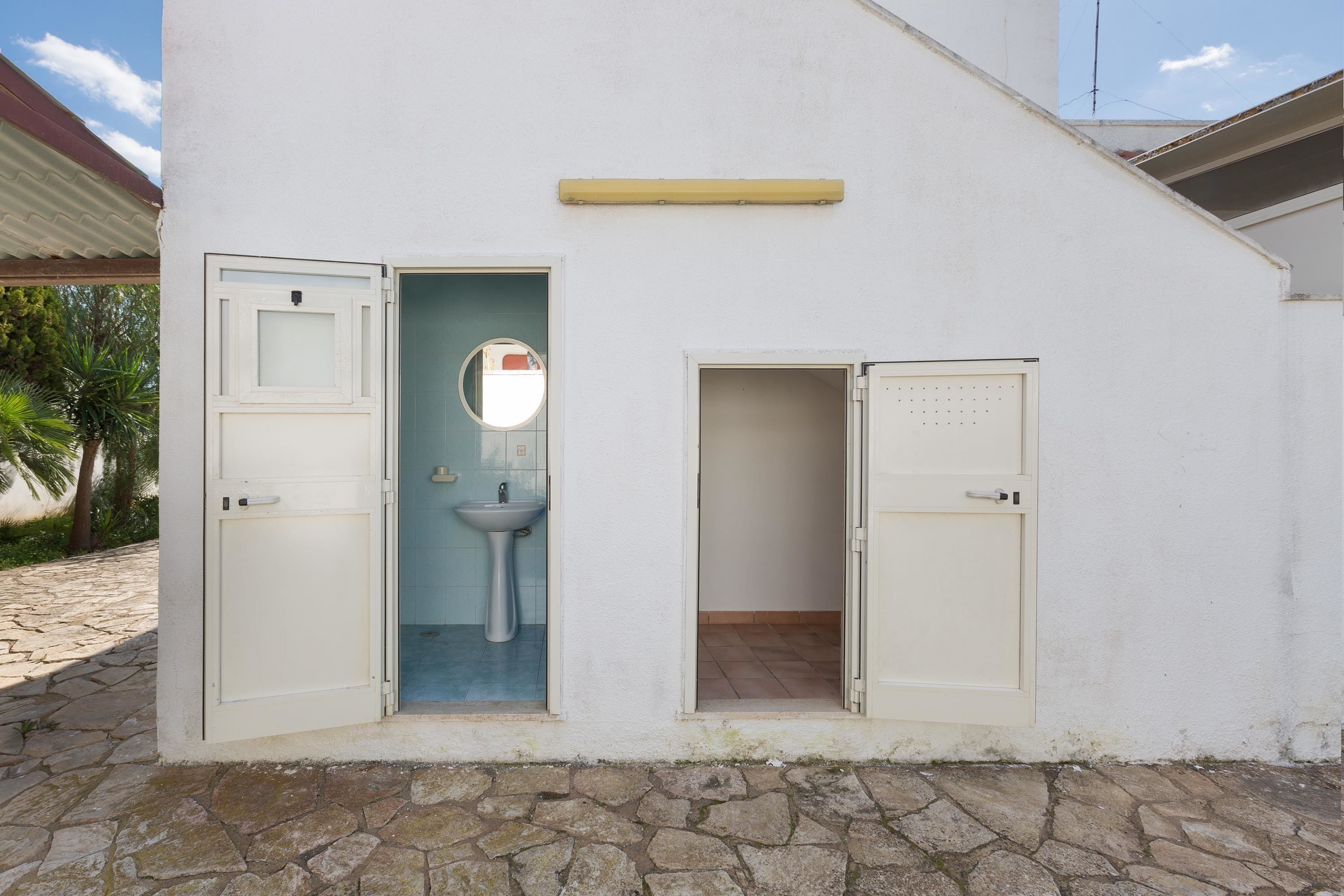 A white building with two open doors, one leading to a bathroom with a sink and mirror, and the other to a small room.