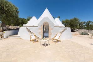 A white, pyramid-shaped building with a stone entrance, two white lounge chairs, and a small table with a bottle and a glass.