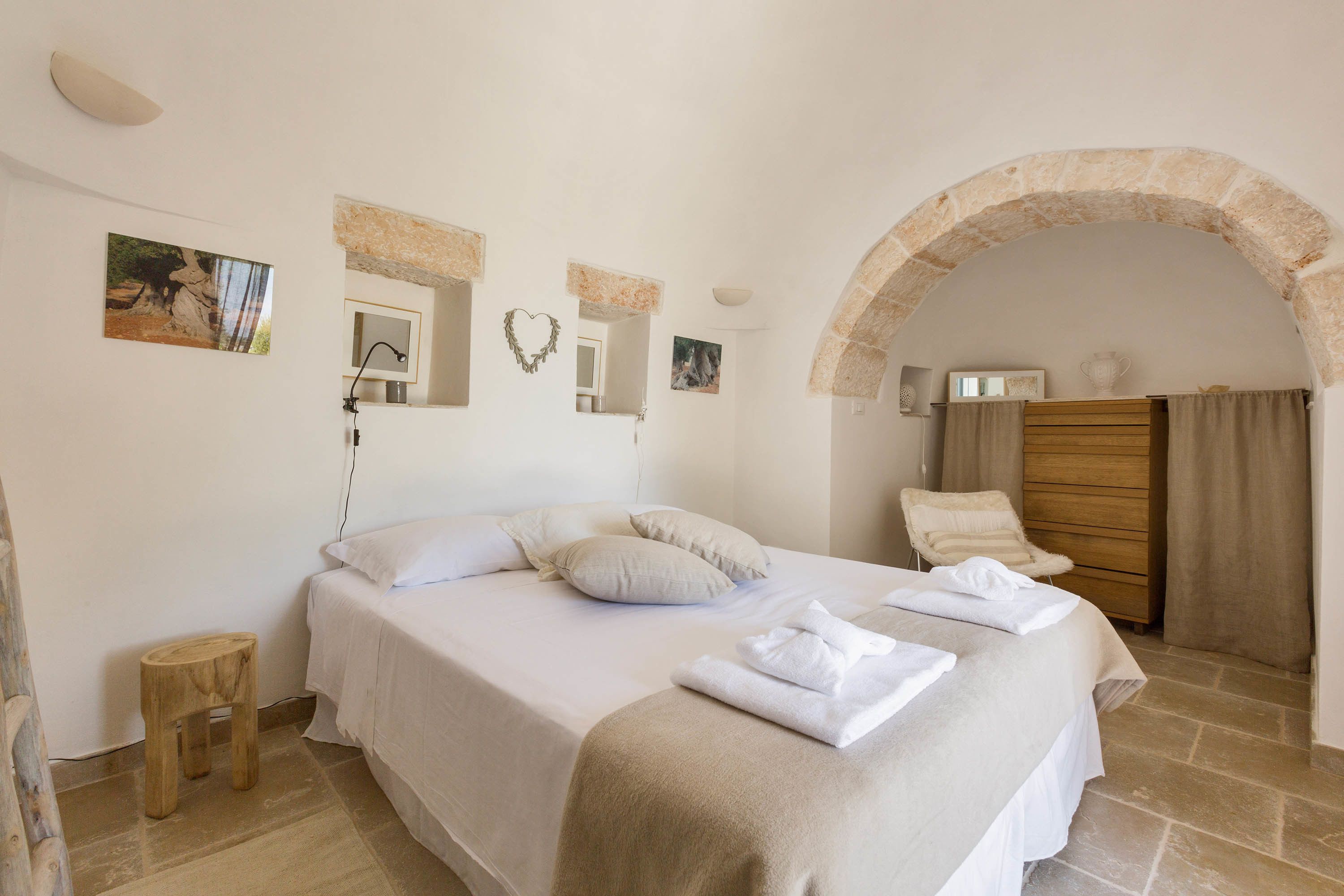 A bedroom with a large bed, a wooden chair, a small stool, and a stone archway.