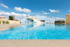 A swimming pool with a clear blue water surface, a covered seating area with blue chairs, and a building with a light blue facade in the background. - Villa Vincenti (Ruffano)