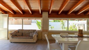 A spacious room with a wooden ceiling, a sofa, a dining table with chairs, and large windows offering a view of greenery outside. - Villetta Giorgina (Porto Cesareo)