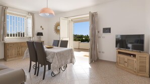 A well-lit room with a dining table, chairs, a TV, and a balcony with curtains. - Villetta di Paulo (Carovigno)