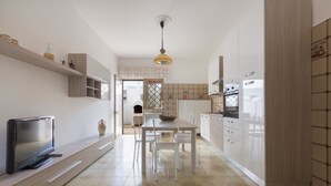 Une cuisine moderne avec des meubles blancs, une table à manger avec des chaises, un téléviseur posé sur un meuble bas et un sol à motifs.