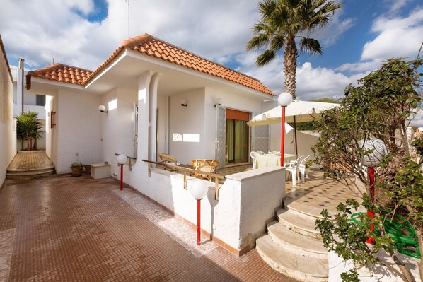 A covered outdoor area with a tiled floor, a palm tree, and a staircase leading to a door.
