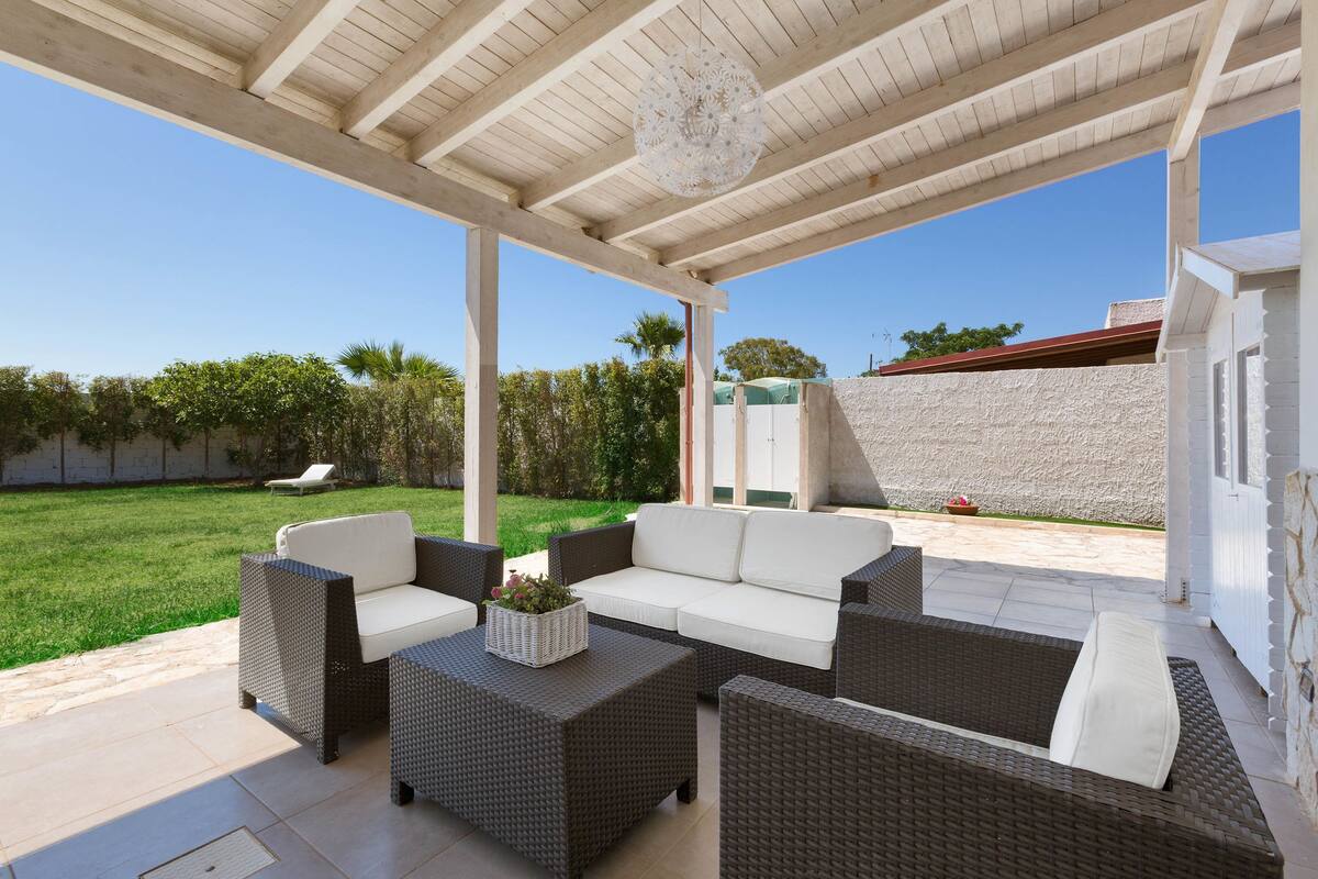 A covered outdoor seating area with wicker furniture and a white sofa, a decorative light fixture, and a view of a green lawn and trees.