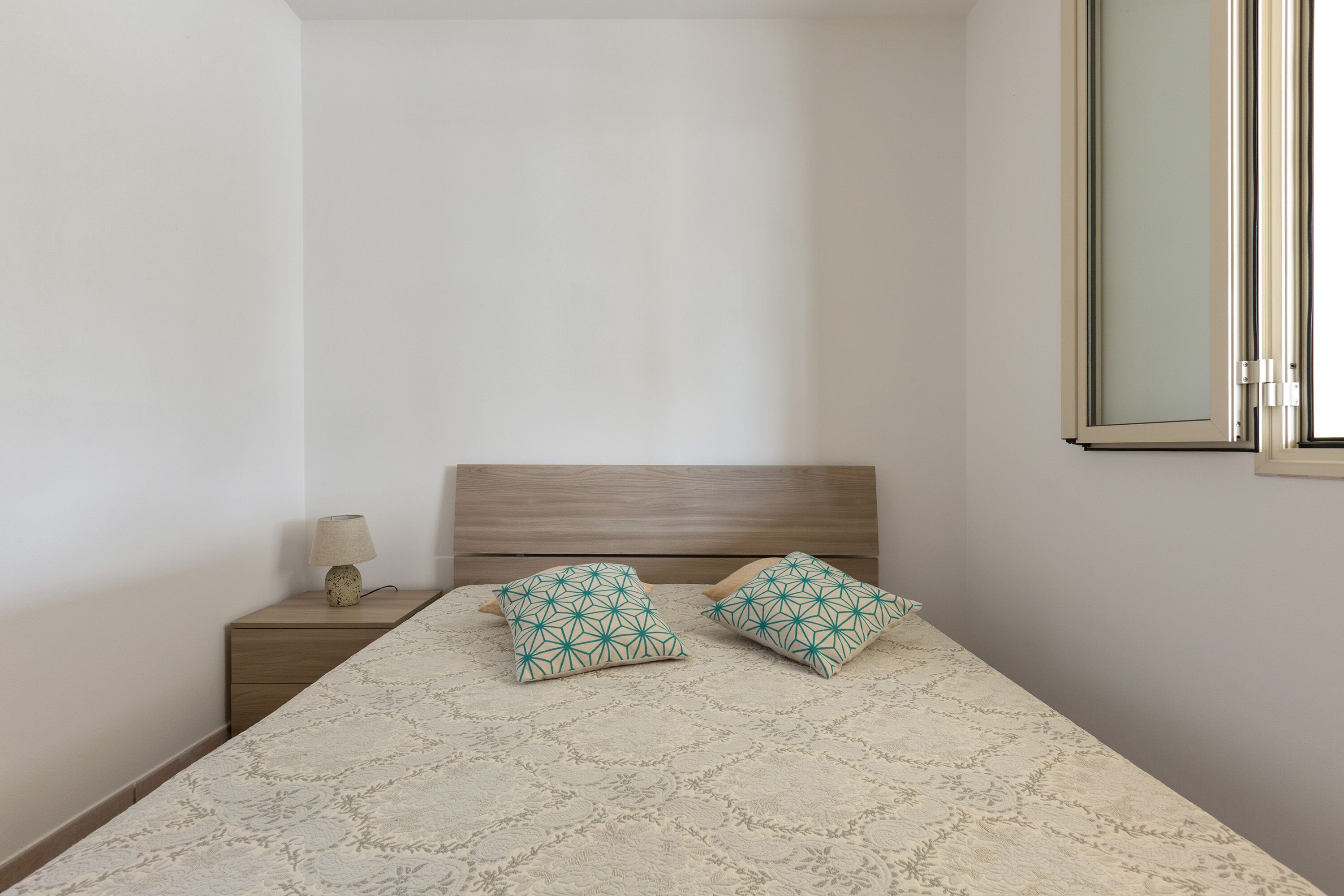 Ein Schlafzimmer mit einem Bett, zwei Kissen, einem Nachttisch, einer Lampe und einem Fenster.