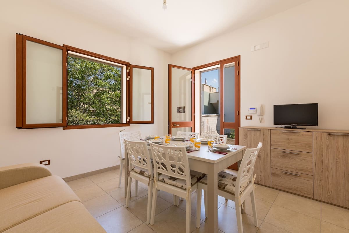 A dining area with a table set for four, white chairs, a television on a wooden cabinet, and a window with a view of trees.