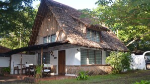 A rustic house with a thatched roof, a wooden porch, and a stone wall. - Diani house (Diani Beach)