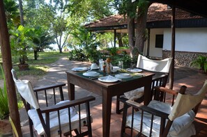 An outdoor dining area with a table set for four, surrounded by chairs, in a tropical setting with trees and a building in the background. - Diani house (Diani Beach)