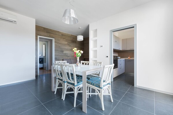 Un espace repas moderne avec une table et des chaises blanches, un vase rempli de fleurs et, au fond, une cuisine.