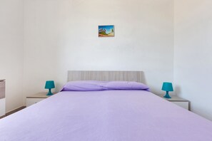 A single bed with a light purple bedspread in a room with white walls. There are two nightstands with teal lamps on either side of the bed.