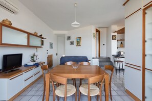 A dining area with a wooden table and chairs, a television on a cabinet, and a kitchen in the background.