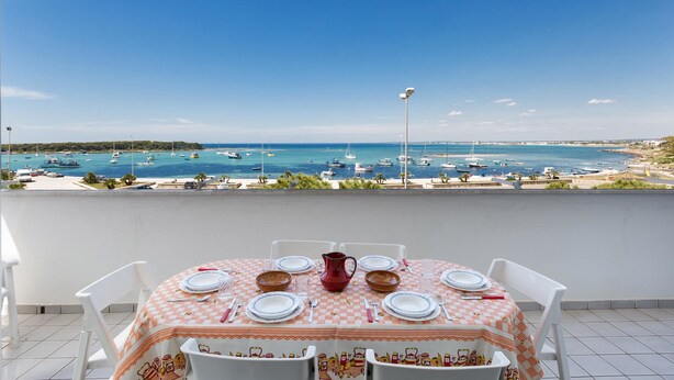 A balcony with a table set for dining, overlooking a marina with boats and a beach. -  909 Appartamento Porticciolo by Barbarhouse (Porto Cesareo)