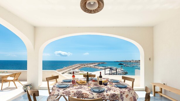 A dining area with a table set for two, overlooking a coastal view with a pier and boats. - Attico del Blu (Castrignano del Capo)