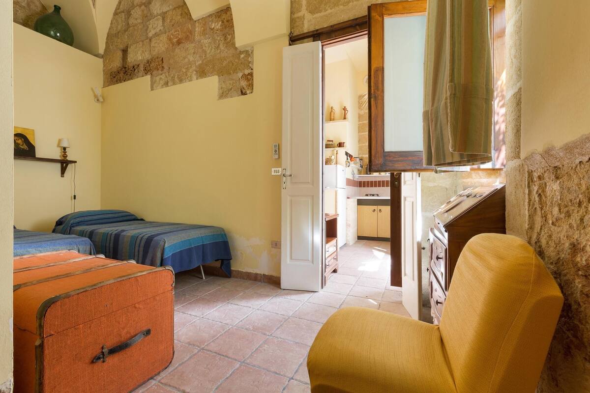 A room with two beds, a suitcase, a chair, a kitchen area, and a stone wall.
