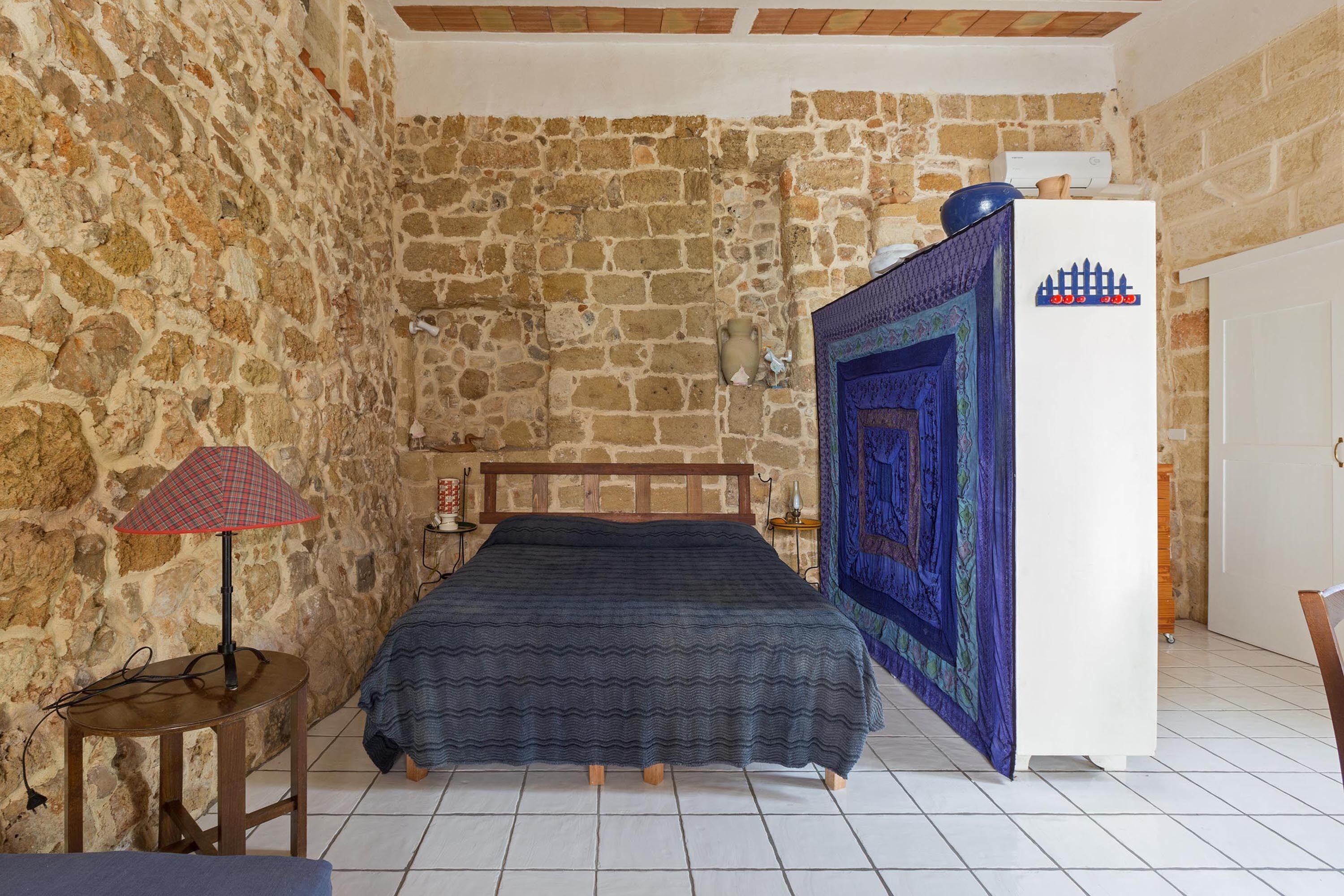 Una camera da letto con parete in pietra, un letto con coperta blu, una lampada e un frigorifero.