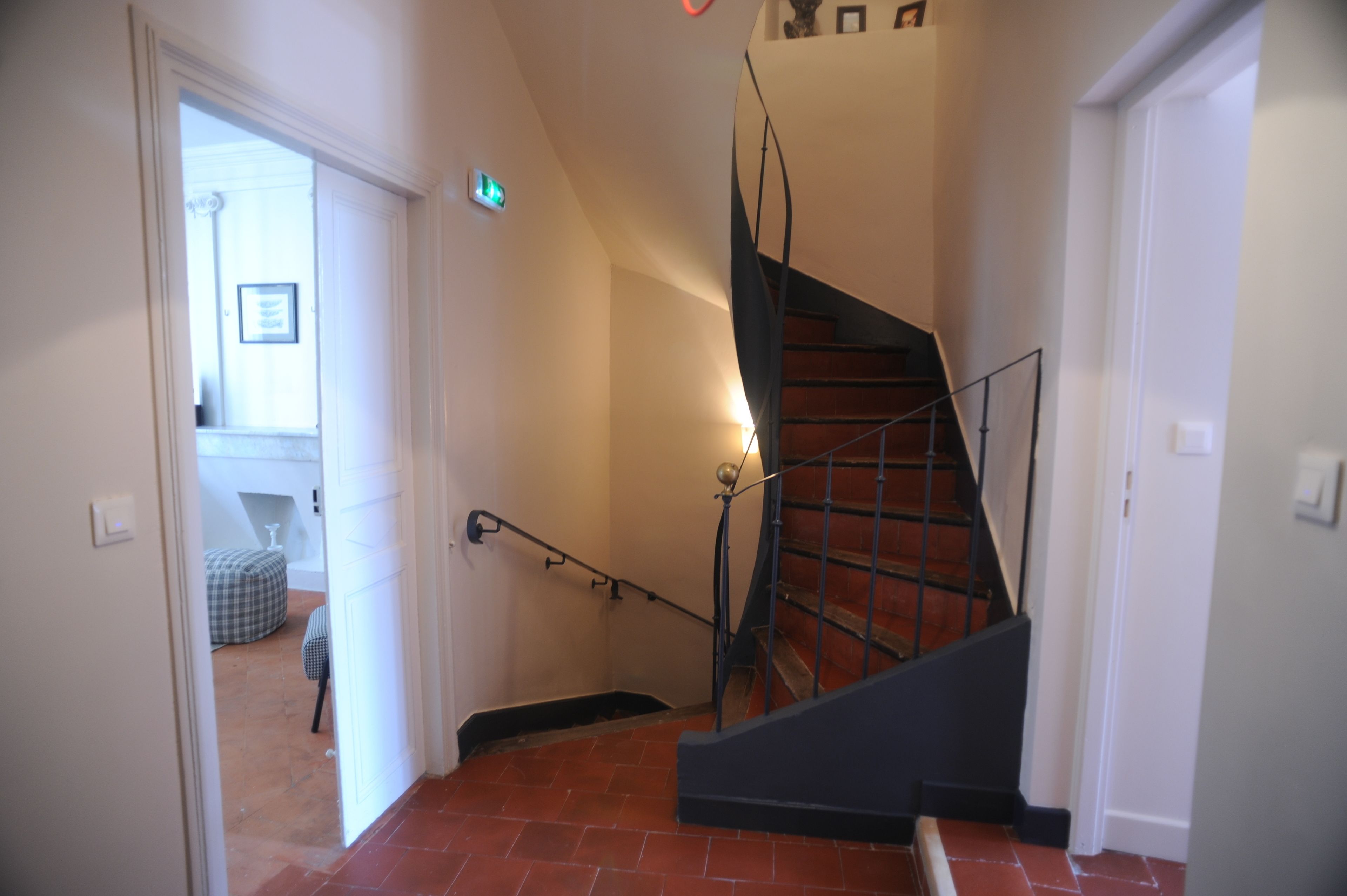 Un escalier avec une main courante noire et un sol carrelé de dalles rouges, menant à une pièce où se trouvent un lit et une chaise.