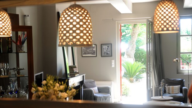 A living room with a fireplace, a coffee table, and a bookshelf. There are two hanging lamps and a window with a view of greenery outside. - Les Chambres d'Andréa (Marseillan)