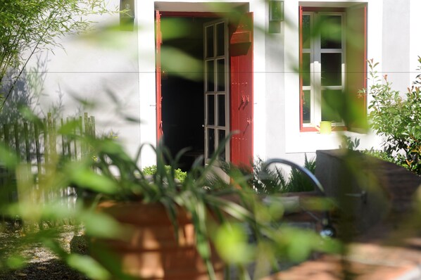 A white building with a red door and window frames, surrounded by greenery.