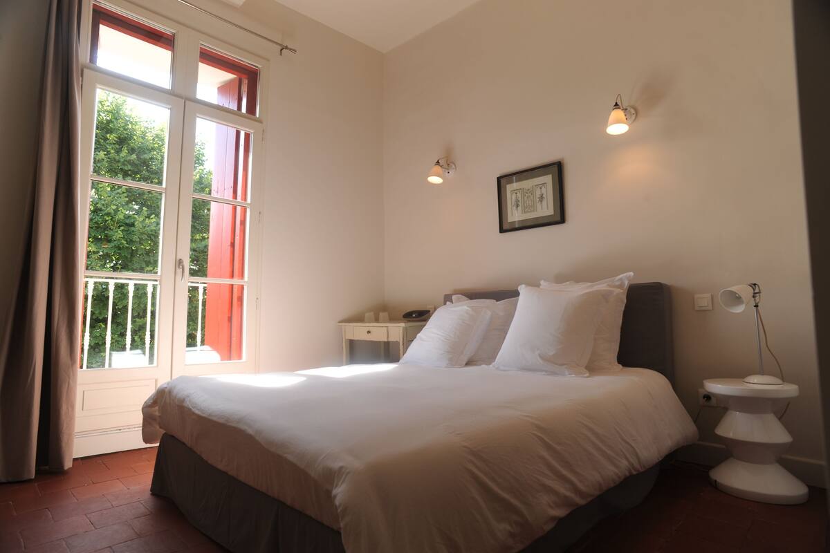 Un dormitorio con cama, mesita de noche, lámpara de noche, ventana con cortinas y un cuadro colgado en la pared.