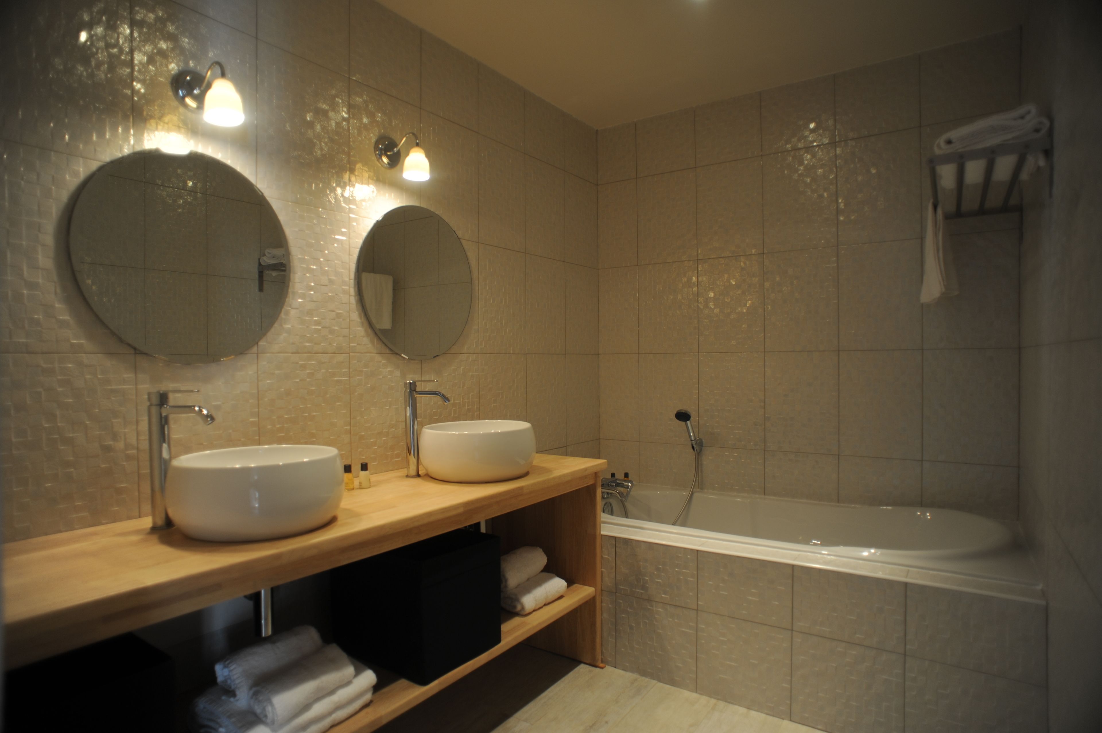 A modern bathroom with two round mirrors, a wooden vanity, a white sink, a bathtub, and a towel rack.