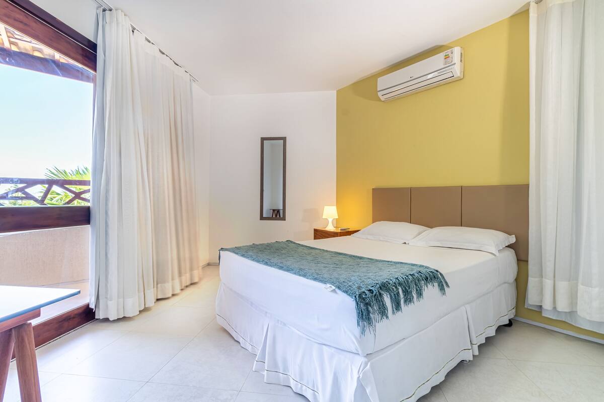 A hotel room with a bed, a nightstand, a lamp, an air conditioner, and a view of the outdoors.