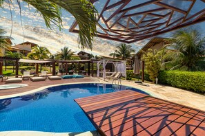 Una piscina con una terraza de madera, sillas de descanso y una zona de estar cubierta.