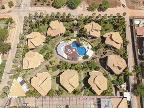 Vista aérea de un complejo turístico con varias construcciones tipo bungalow, una piscina central y palmeras a su alrededor.