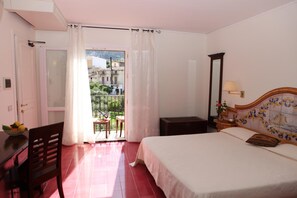 A bedroom with a bed, a desk, a chair, a balcony with a view of buildings, and a window with curtains. - Hotel Torre Bennistra (Castellammare del Golfo)