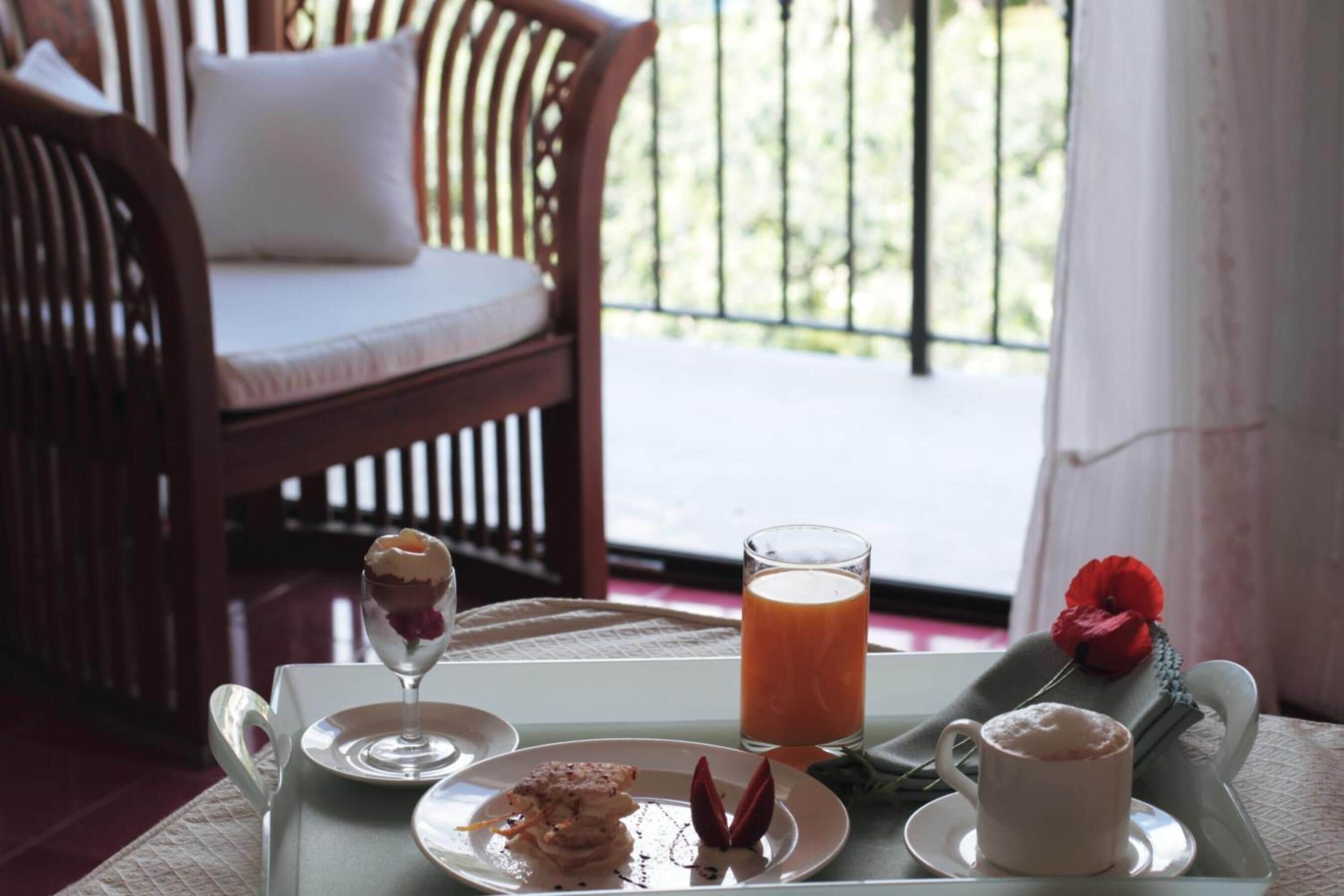 A dining table with a dessert, a glass of juice, and a cup of coffee, set on a balcony with a view of greenery.