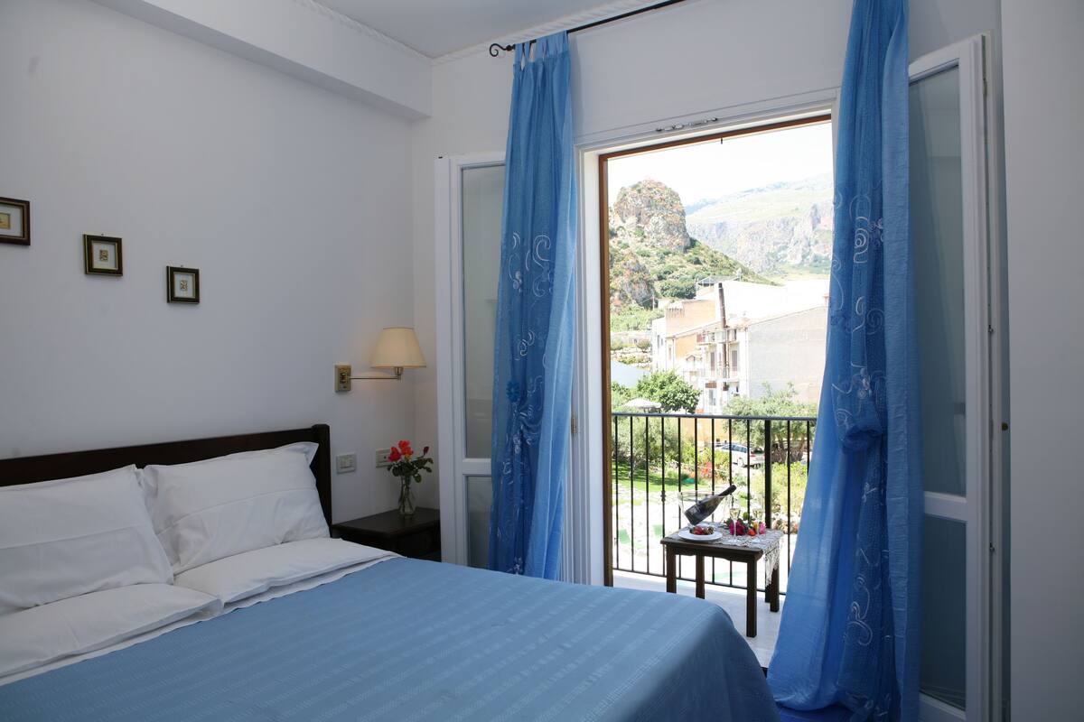 A hotel room with a bed, a nightstand with a lamp, a small table with a vase of flowers, and a balcony with a view of the city and mountains.