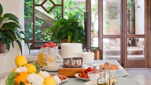 Un bufé de desayuno con diversas opciones de comida y bebidas, como frutas, quesos y pan, dispuesto sobre una mesa en una habitación con ventanas amplias y vistas a la vegetación exterior.