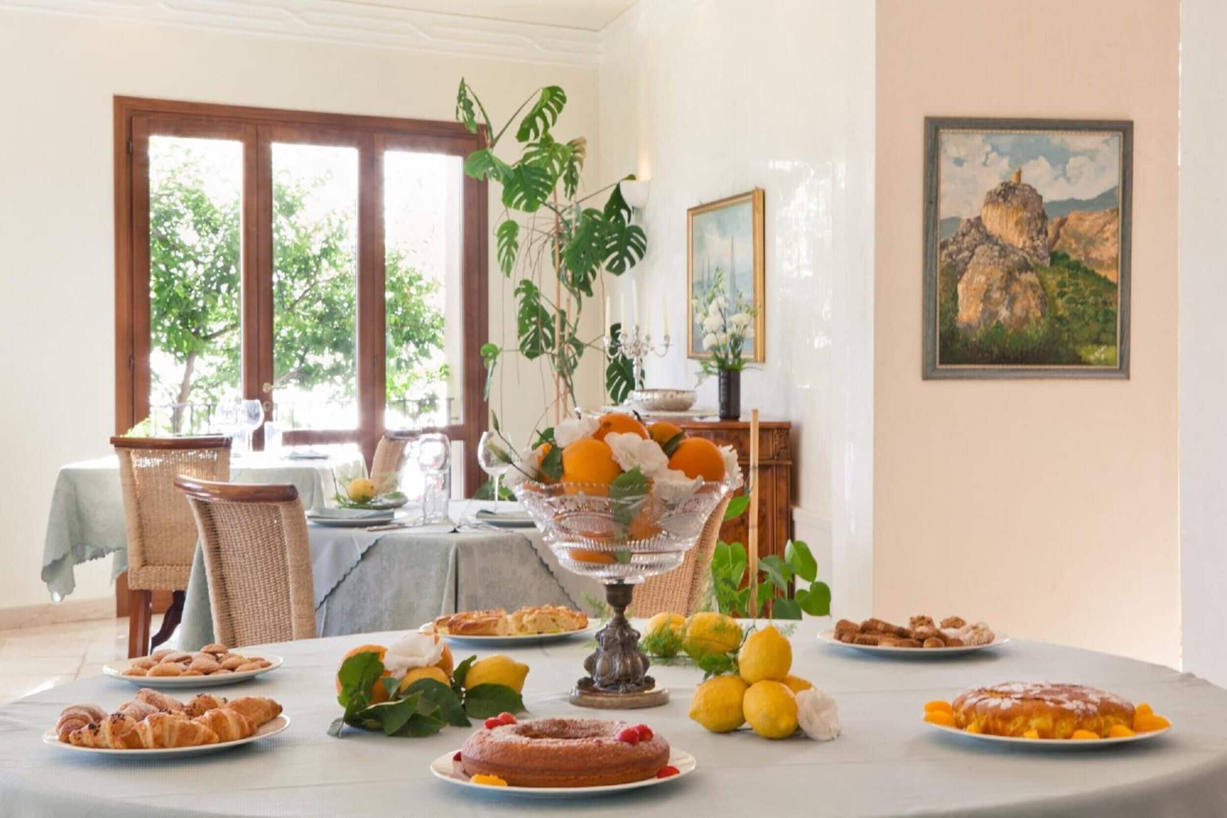 A dining table set with various pastries and fruits, surrounded by wicker chairs and a large window with a view of greenery.